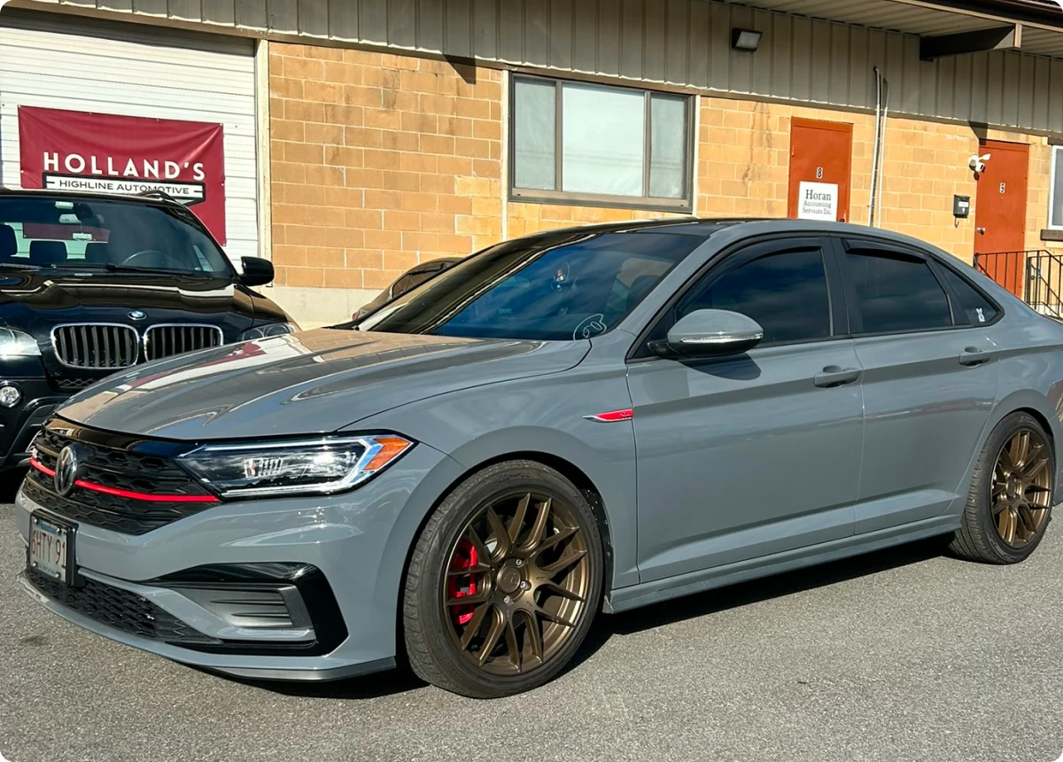 A gray Volkswagen sedan with bronze wheels and red brake calipers is parked outside a tan brick building with a Hollands Highline Automotive sign, showcasing expert Volkswagen repair & services in Bridgewater.