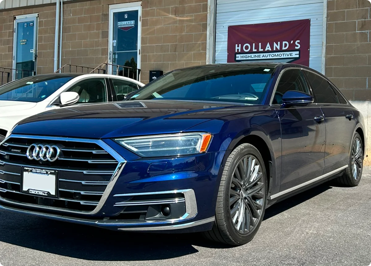 A blue Audi sedan, serviced by an Audi expert, is parked outside a building with a “Holland’s Highline Automotive” sign. Another white car is partially visible beside it, and stairs lead to doors in the background.
