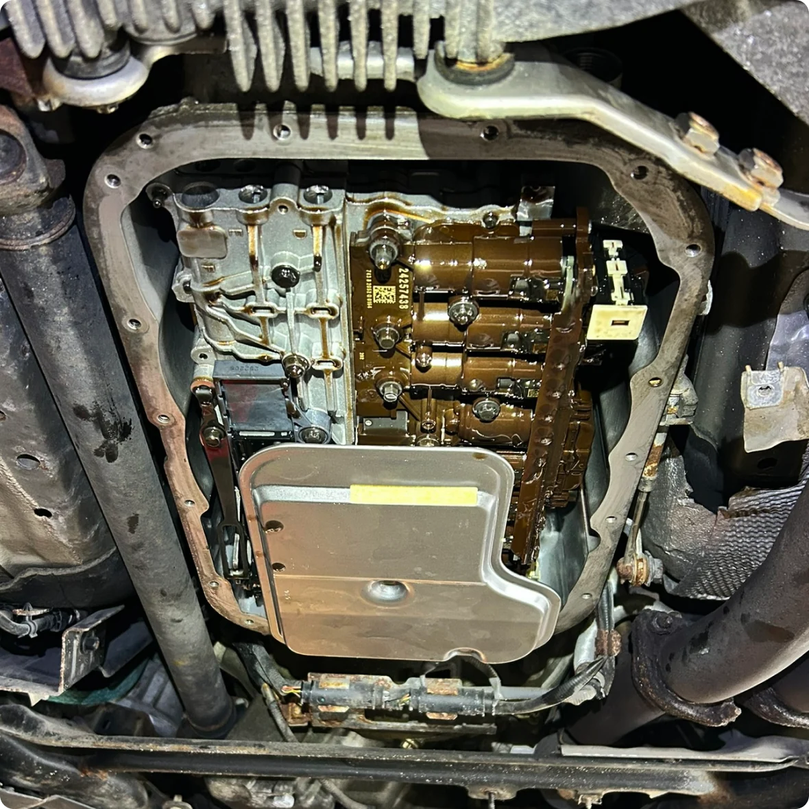 View of the underside of a vehicle during transmission repair and replacement, showing an exposed automatic transmission with the oil pan removed, internal components coated in fluid, and various surrounding metal parts and pipes.