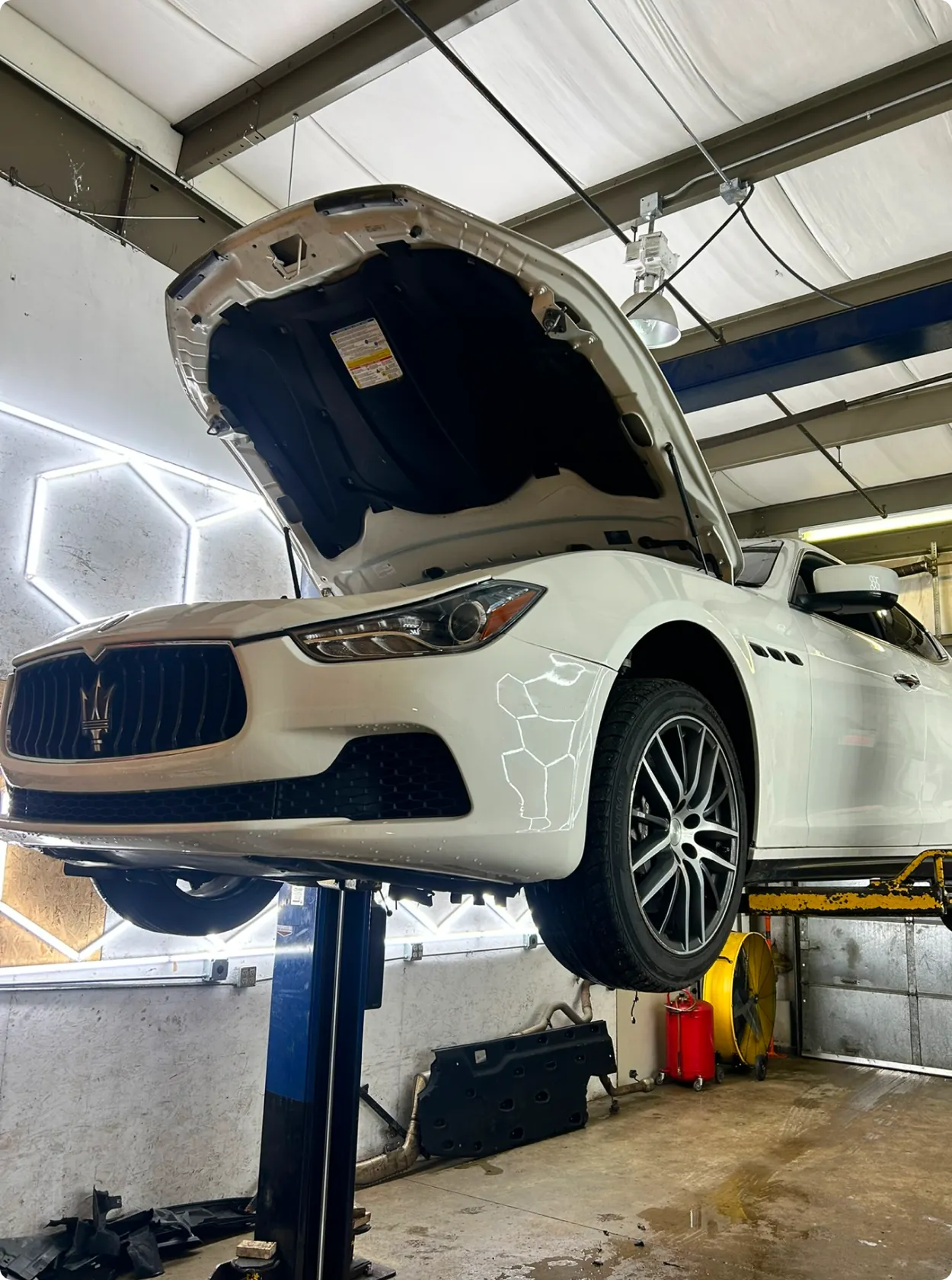 A white Maserati with its hood open is elevated on a car lift in an auto repair shop, showcasing tools, equipment, and overhead lighting—highlighting european auto repair in Bridgewater.