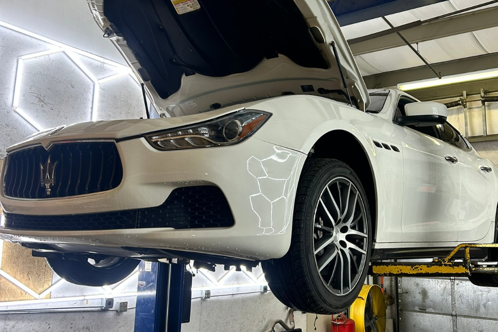 European Auto Repair in Bridgewater, MA at Holland's Highline Automotive. A white sports car with its hood open is lifted on a hydraulic car lift in a garage, where a European auto repair mechanic works. Neon lights and tools fill the background, and white marker outlines are drawn on the car’s front bumper.