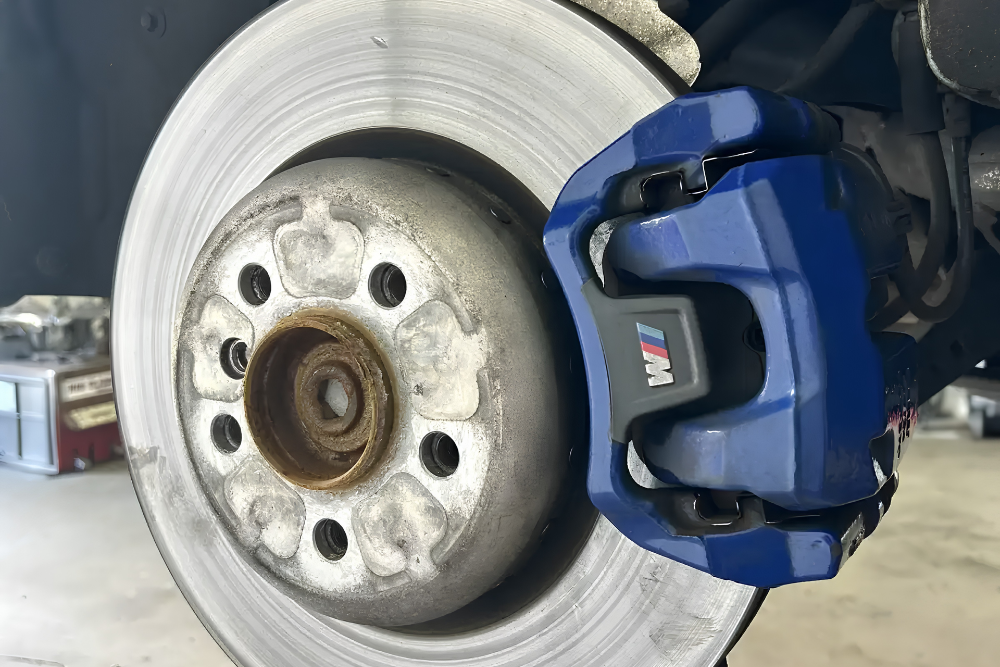 Brake Repair in Bridgewater, MA At Holland’s Highline Automotive. Close-up of a cars disc brake system, featuring a silver brake rotor with signs of rust and wear, and a blue brake caliper with an M logo, likely indicating a performance part. The background shows a garage setting.