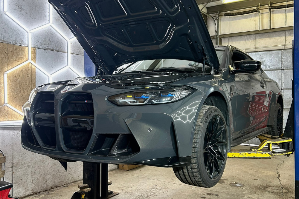 Vehicle Inspection in Bridgewater, MA at Holland's Highline Automotive. A gray sports car with its hood open is lifted on a car lift inside a garage with illuminated hexagonal wall panels, ready for a thorough inspection.